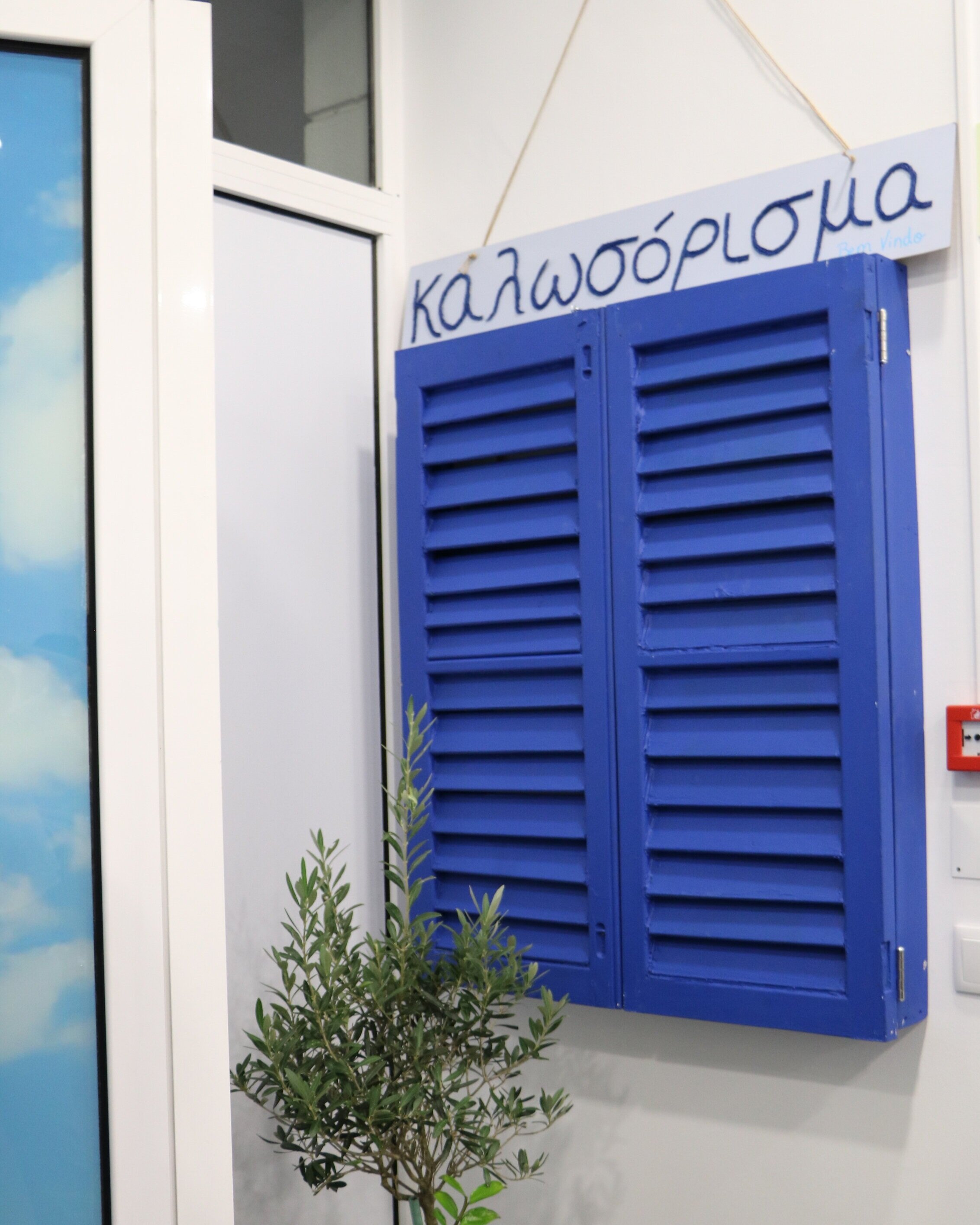 Placa de boas-vindas em grego, junto a uma janela azul decorativa